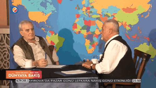 two men are sitting at a table with a map of the world in the background