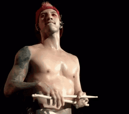a shirtless man wearing a red hat is holding a pair of drumsticks .