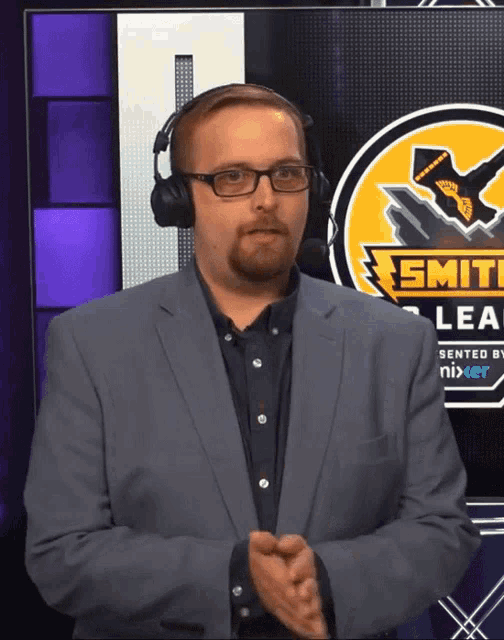 a man wearing a suit and headphones stands in front of a smite league logo