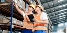 two workers are standing in a warehouse looking at boxes on shelves .