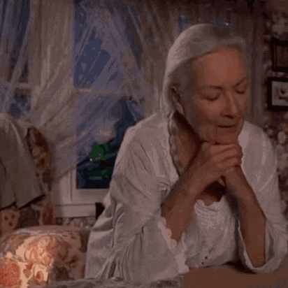 an elderly woman sits in front of a window with her hands folded in prayer