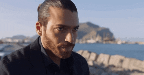 a man with long hair and a beard looks at the camera in front of a body of water