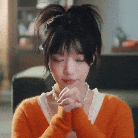 a woman in an orange sweater and pearls is praying with her eyes closed .