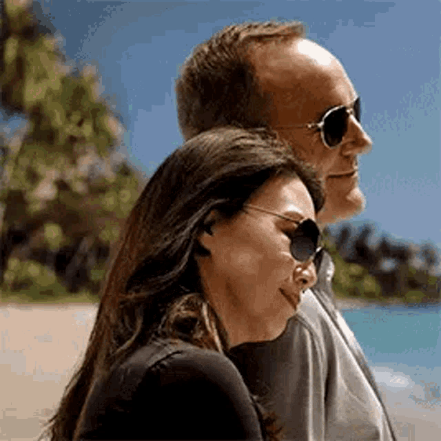 a man and a woman are standing next to each other on a beach . the woman is wearing sunglasses .