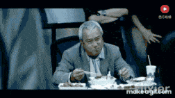 a man is sitting at a table with a spoon in his mouth and a play button on the bottom right