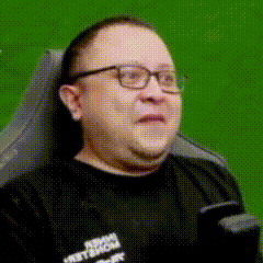 a man wearing glasses and a black shirt is sitting in a chair with a green screen behind him .