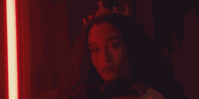 a woman with a crown on her head is sitting in a dark room with a red light behind her .