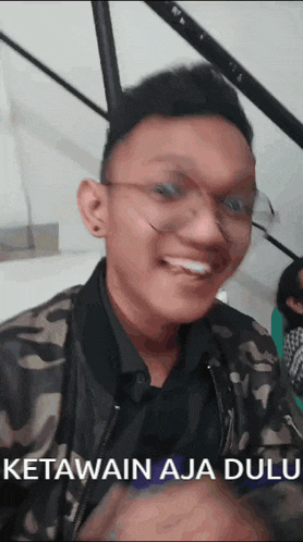 a man wearing glasses and a camouflage jacket is smiling with the words ketawain aja dulu above him