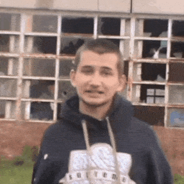 a young man wearing a hoodie is standing in front of a building with broken windows .
