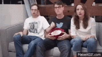 three people are sitting on a couch eating popcorn while watching a movie .