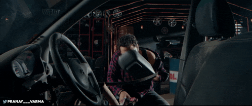 a man in a plaid shirt is sitting in the driver 's seat of a car with the words pranay varma below him