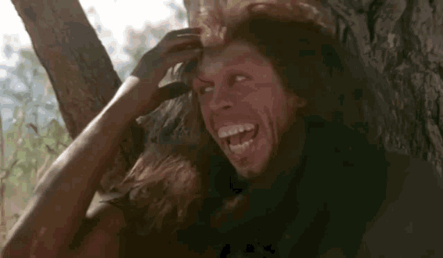a man with long hair is laughing while leaning against a tree trunk .