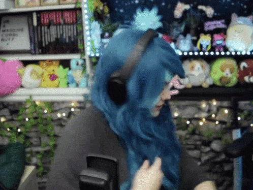 a person with blue hair wearing headphones in front of a shelf full of stuffed animals