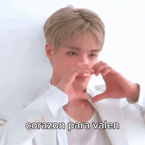 a young man in a white shirt is making a heart shape with his hands and the words corazon para valen below him .