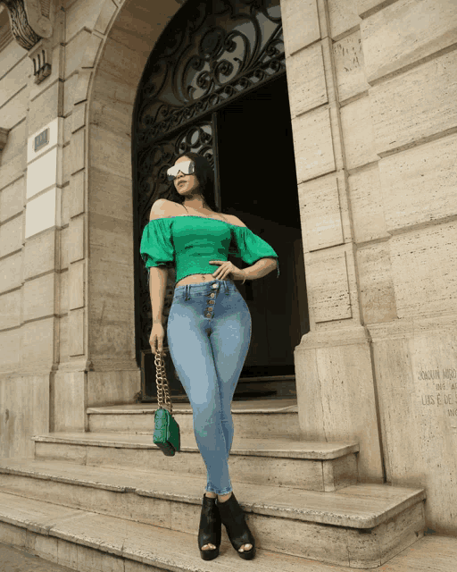a woman wearing a green off the shoulder top and jeans stands in front of a building