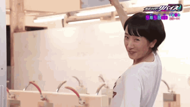 a woman in a white t-shirt is standing in front of a wall that says ' kamen rider ' on it