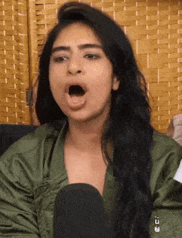 a woman in a green jacket is talking into a microphone with her mouth open .