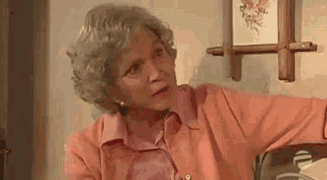 a woman with gray hair and a pink shirt is sitting at a table in front of a wall with a picture on it .