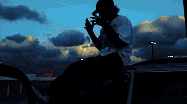 a silhouette of a person sitting on the roof of a car smoking a cigarette