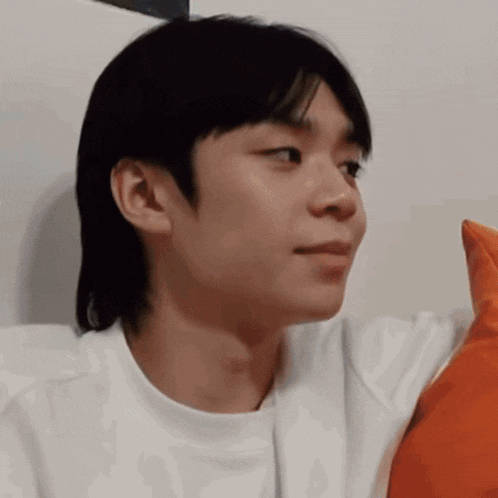 a young man is sitting on a couch with an orange pillow and looking at the camera .