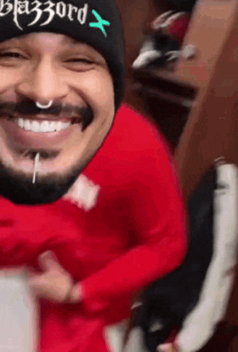 a man with a beard wearing a beanie and a red sweatshirt is smiling .