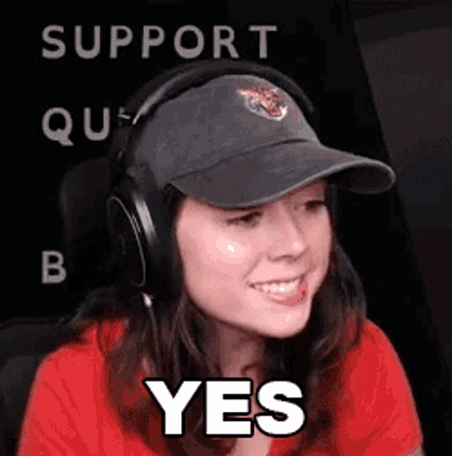 a woman wearing headphones and a baseball cap is smiling and saying yes .
