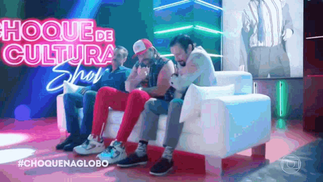 three men are sitting on a white couch in front of a sign that says hoque de cultura show