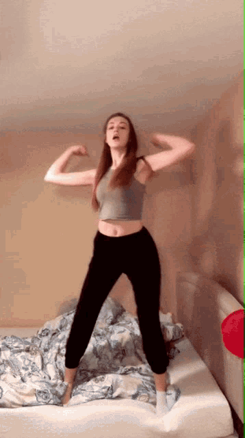 a woman in a grey tank top and black pants is standing on a bed