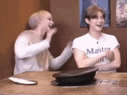 two young men are sitting at a table laughing . one of the men is wearing a white shirt that says master .