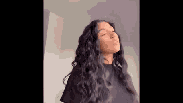 a woman with long curly hair is wearing a black shirt and standing in front of a wall .