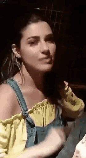 a woman in a yellow off the shoulder top and blue overalls is making a funny face