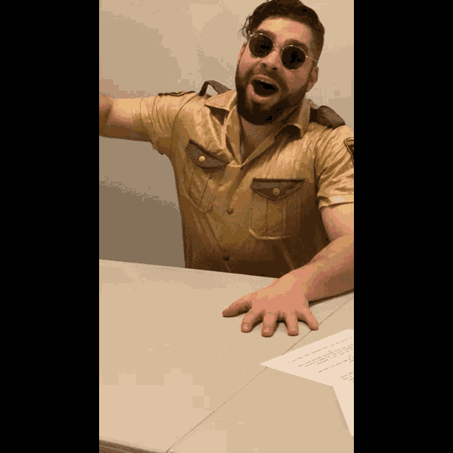 a man with a beard wearing sunglasses and a gold shirt is sitting at a table