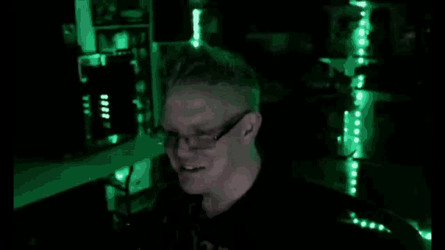 a man wearing glasses is smiling in a dark room with green lights behind him .