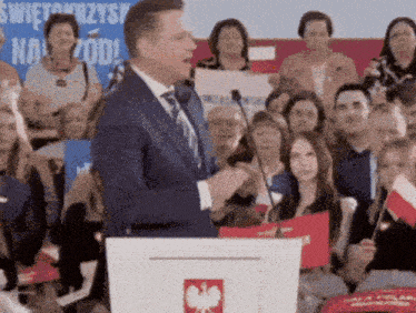 a man stands at a podium in front of a crowd with a sign in the background that says świętokrzysy