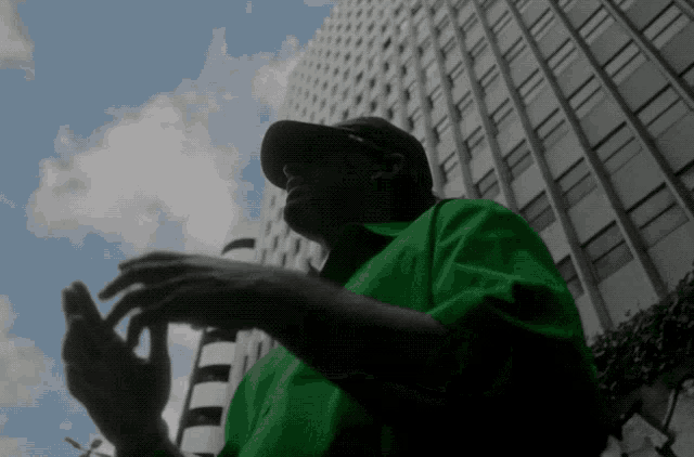 a man in a green shirt and hat stands in front of a tall building