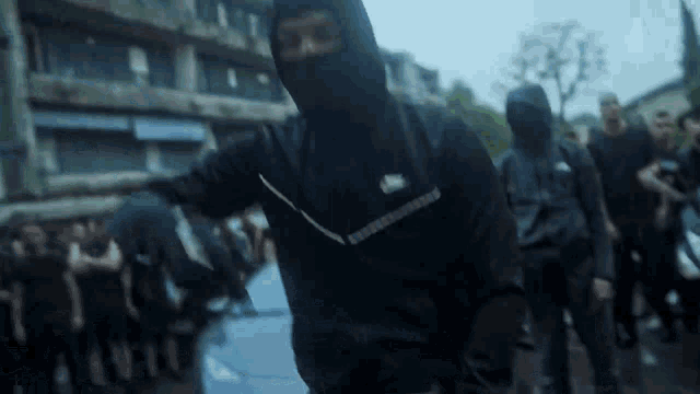 a man in a black hoodie is standing in front of a crowd of people wearing masks .