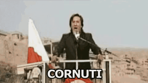 a man in a suit is standing in front of a microphone with the word cornuti written on the screen .