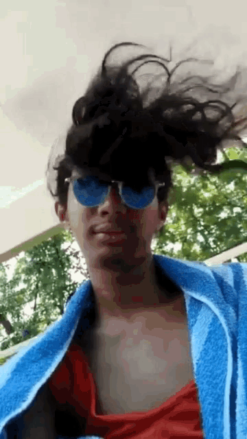 a man wearing sunglasses and a blue towel looks at the camera with his hair blowing in the wind