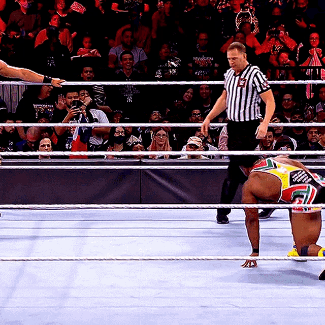 a referee stands in a wrestling ring with a wrestler kneeling down in front of a crowd