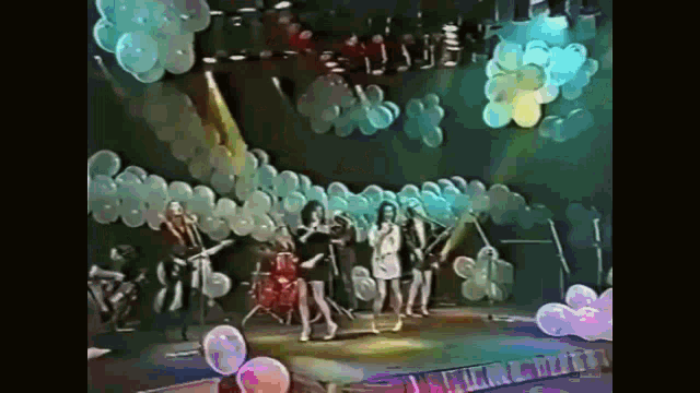 a group of women are dancing on a stage with balloons hanging from the ceiling .