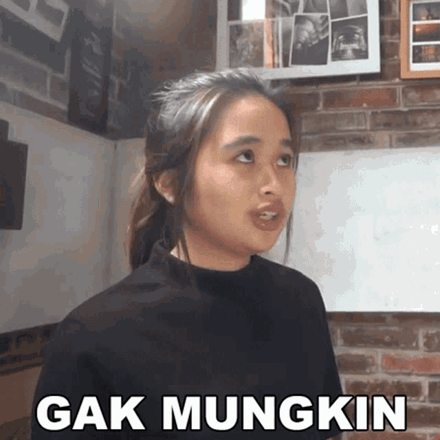 a woman in a black shirt is standing in front of a brick wall with the words " gak mungkin " written on it