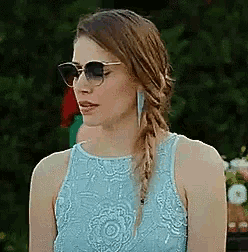 a woman wearing sunglasses and a light blue dress is standing in front of a bush .