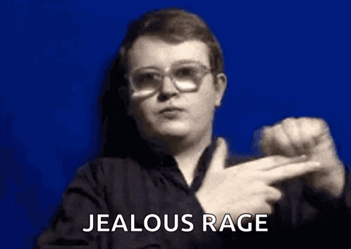 a man wearing glasses is pointing at the camera and saying `` jealous rage '' in sign language .