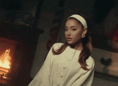 ariana grande is standing in front of a fireplace wearing a white dress .