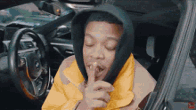 a man in a hooded jacket is sitting in a car with his hand on his mouth .
