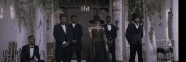 a woman in a black dress is standing in front of a group of men in tuxedos and hats .