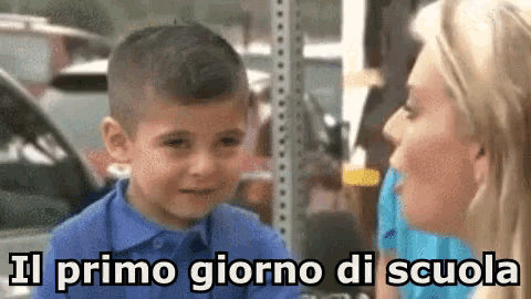 a woman is talking to a young boy who is wearing a blue shirt with the words il primo giorno di scuola written on it