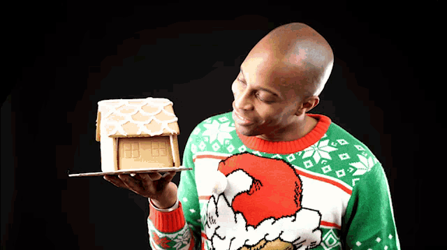 a man in a grinch sweater holds a gingerbread house