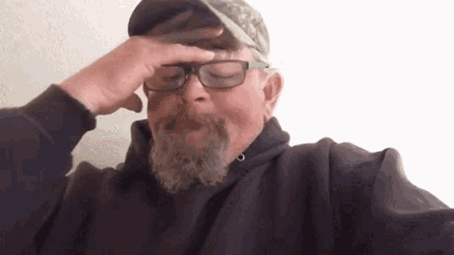 a man with a beard and glasses is wearing a hat and covering his forehead with his hand .