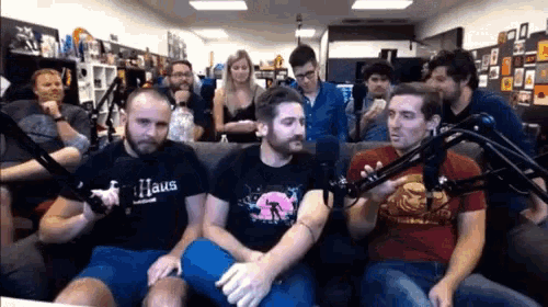 a man wearing a haus shirt sits in a room with other people